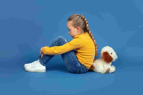Child sitting with knees drawn up beside a stuffed dog against a plain backdrop